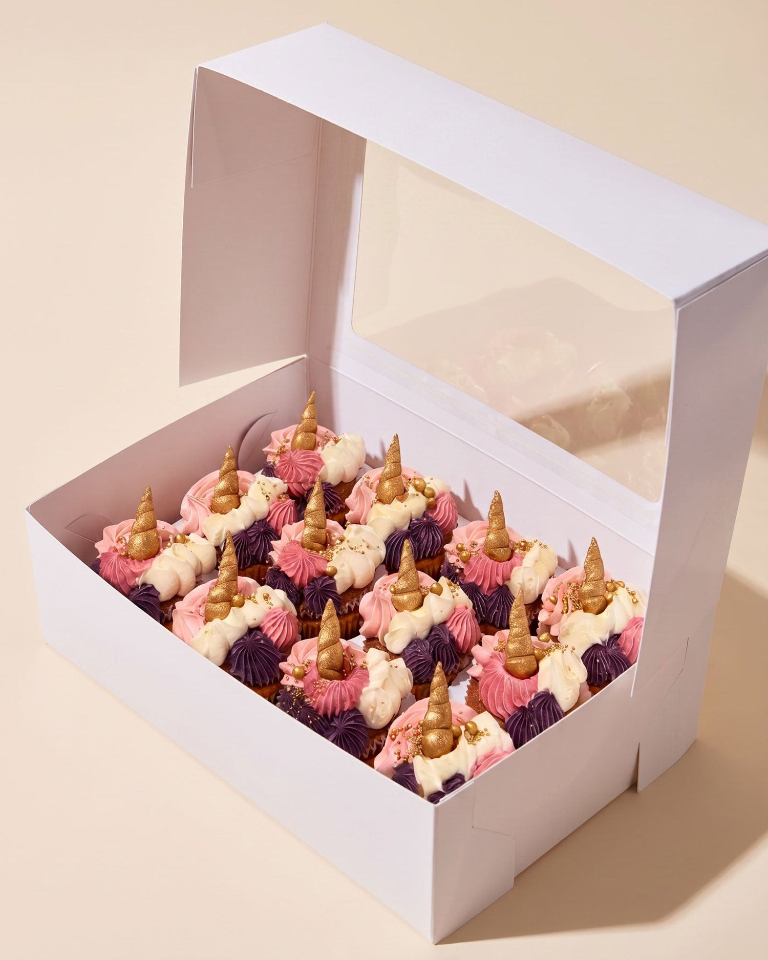 A white bakery box with a clear window lid, filled with decorated cupcakes topped with pink and white frosting, purple drizzle, and gold horn candies, on a warm beige background.