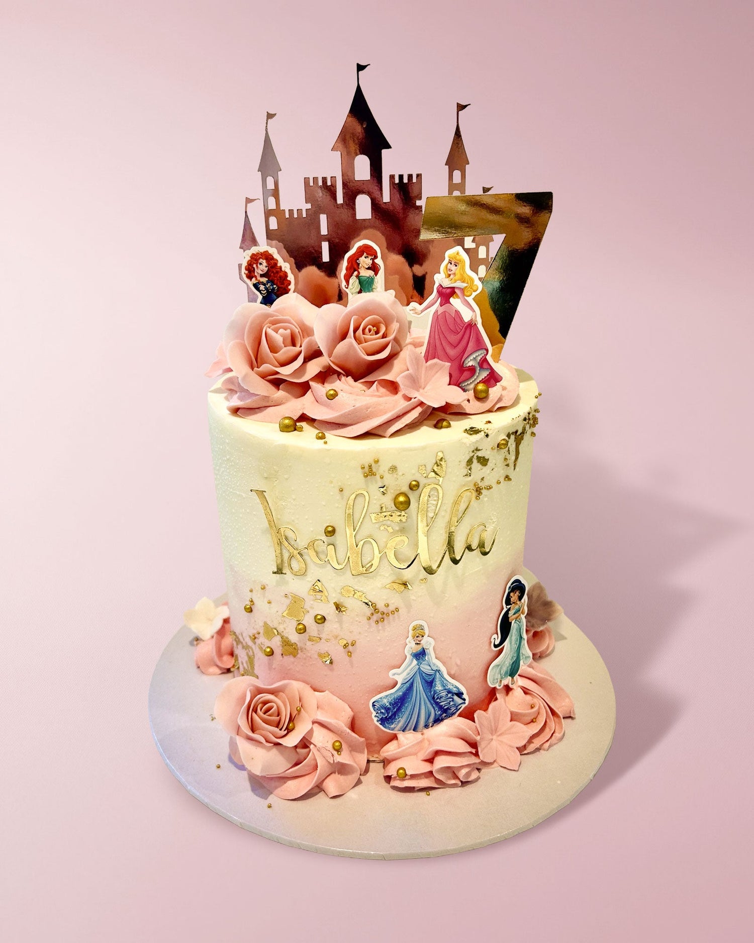 A custom decorated birthday cake with pink frosting roses, gold leaf details, and princess toppers, topped with a castle silhouette on a light pink background.