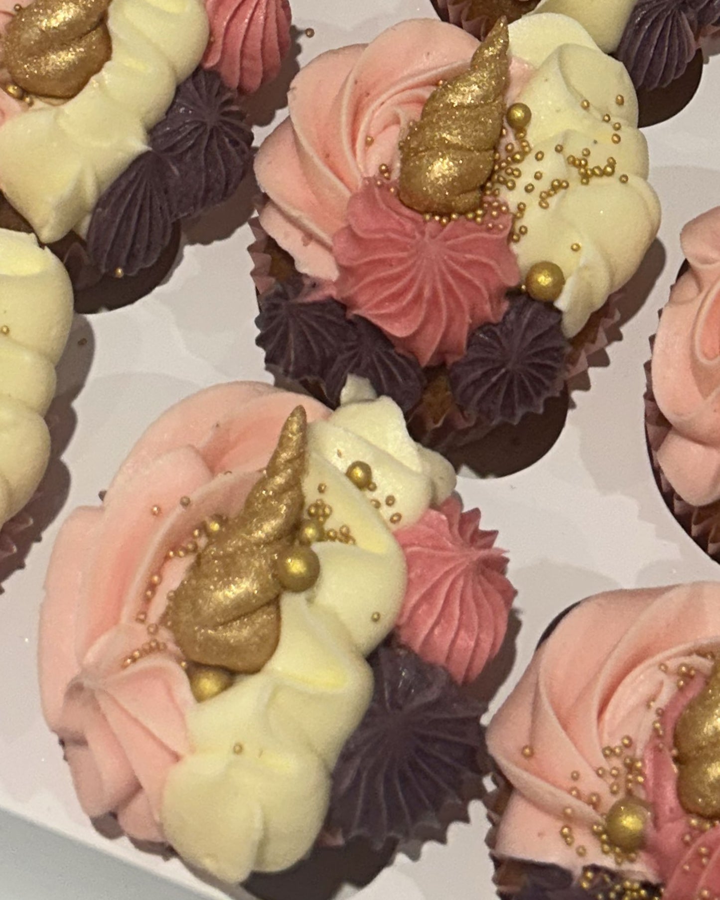 Close-up of decorated cupcakes topped with pink and cream frosting, dark purple rosettes, and a gold foil horn accent with gold sprinkles.