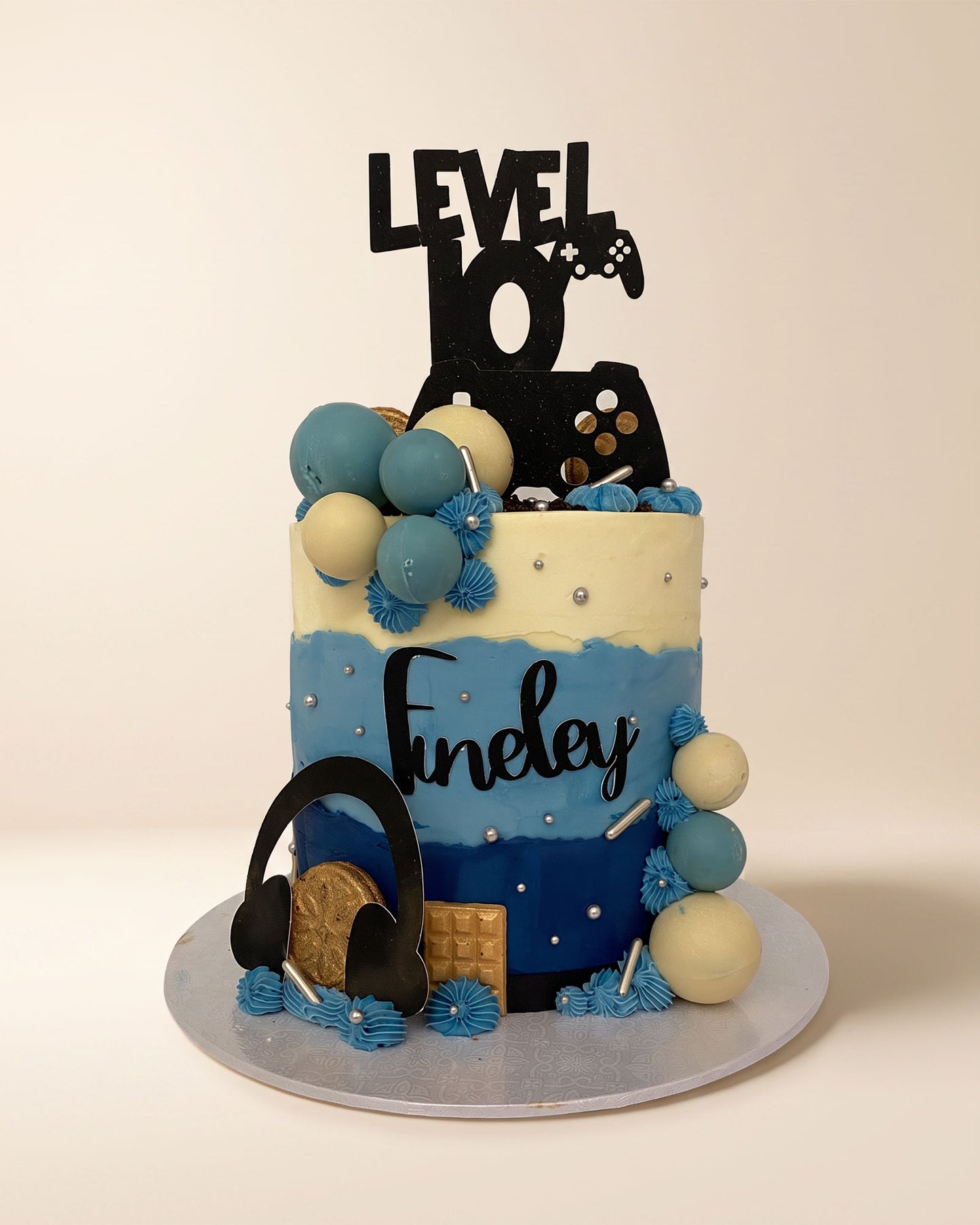 A custom tiered birthday cake decorated with blue and cream frosting, game controller graphics, a black 'LEVEL 10' topper, and a black 'Finley' script name, displayed on a light beige background.