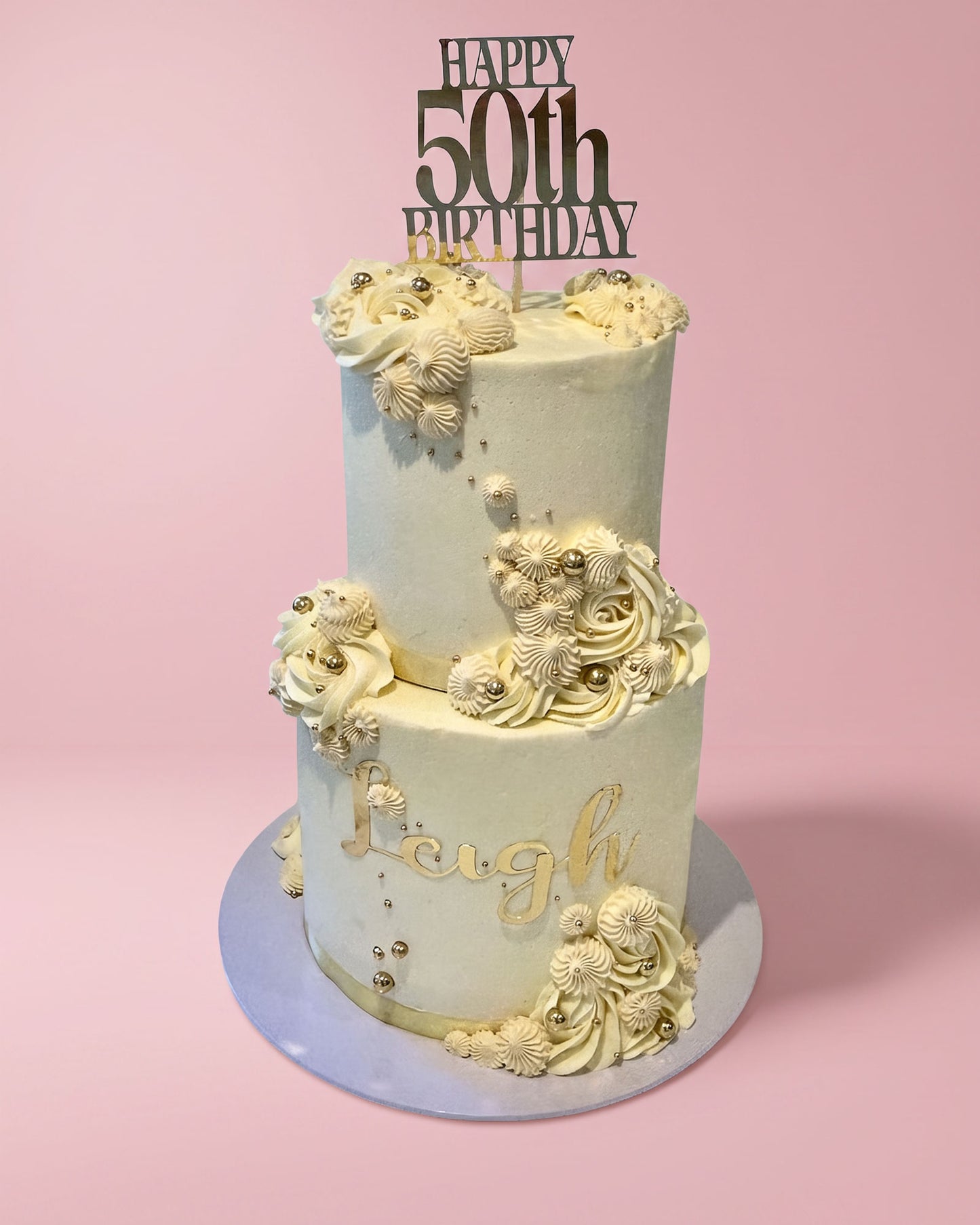 A two-tier frosted birthday cake with cream-colored icing, gold script lettering, and decorative 3D floral and shell-like embellishments, topped with a black 'HAPPY 50th BIRTHDAY' topper, set against a solid pink background.