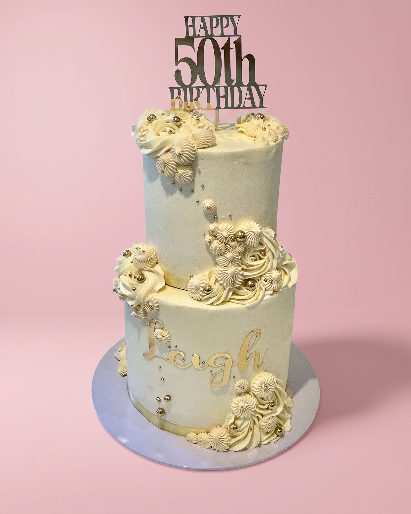 A two-tier frosted birthday cake with cream-colored icing, gold script lettering, and decorative 3D floral and shell-like embellishments, topped with a black 'HAPPY 50th BIRTHDAY' topper, set against a solid pink background.