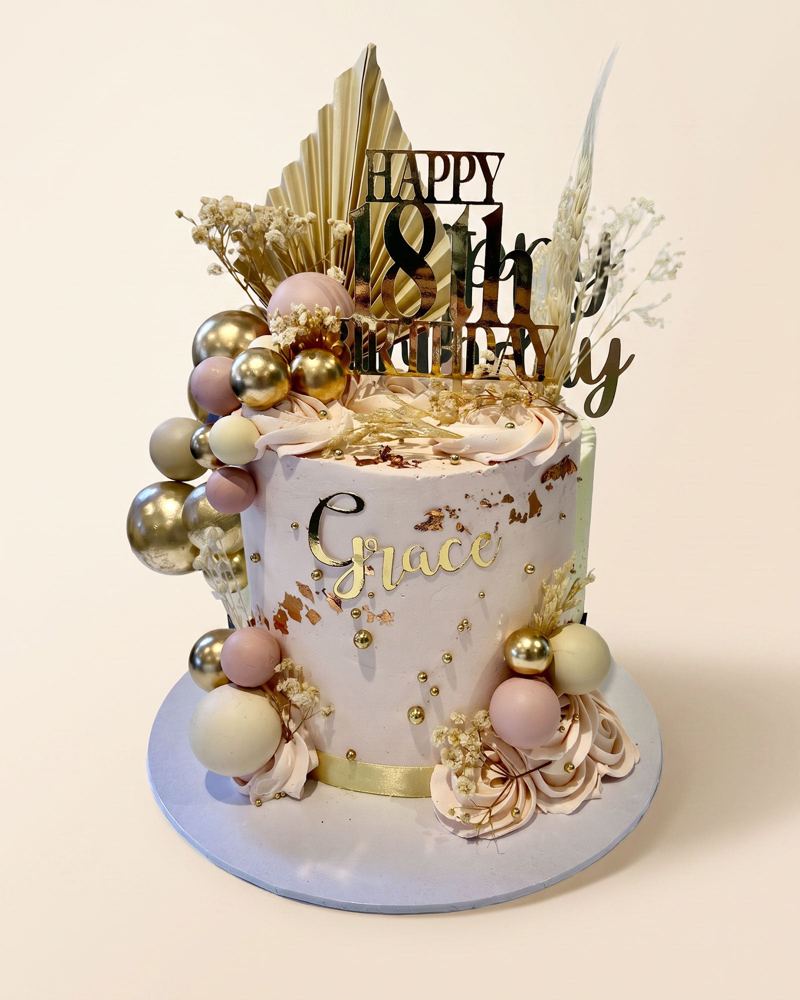 A custom decorated birthday cake with a pale pink and white base, gold foil details, and gold, pink, and white decorative spheres. The cake is topped with a gold 'HAPPY 18th Birthday' topper and dried botanical elements, and stands on a round light blue plate against a solid cream background.