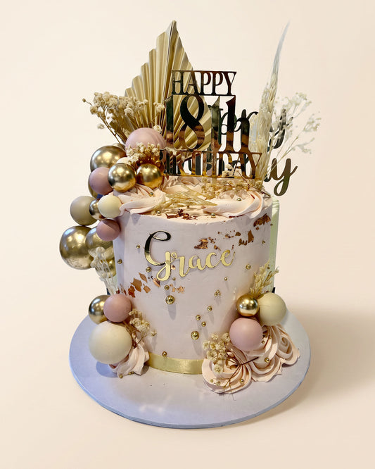 A custom decorated birthday cake with a pale pink and white base, gold foil details, and gold, pink, and white decorative spheres. The cake is topped with a gold 'HAPPY 18th Birthday' topper and dried botanical elements, and stands on a round light blue plate against a solid cream background.