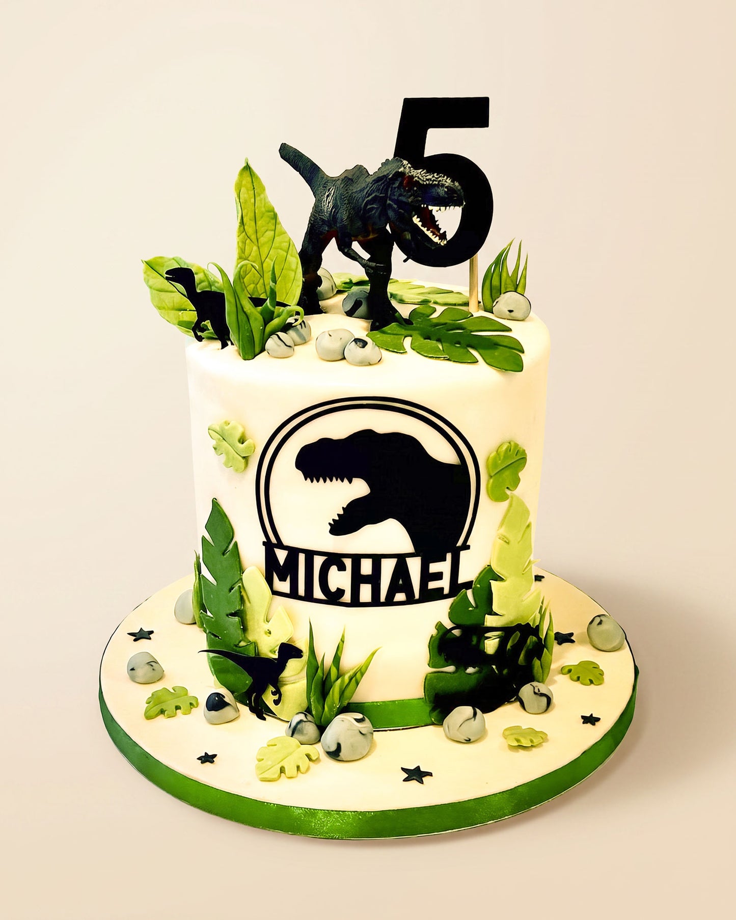 Dinosaur-themed fondant birthday cake with black T-Rex silhouette, green leaves, faux rocks and a personalised number topper, handmade by Daisy Mud Pie in Brentwood, Essex