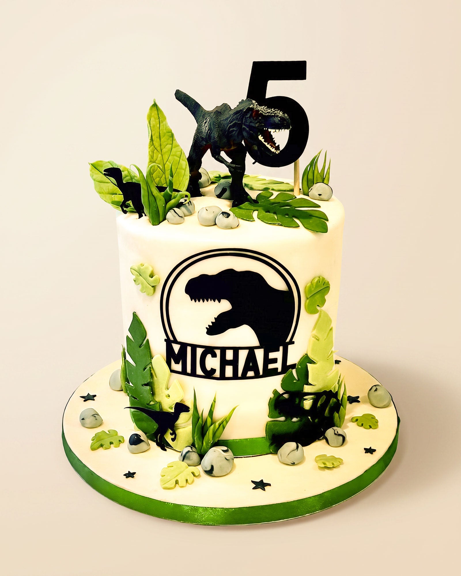 Dinosaur-themed fondant birthday cake with black T-Rex silhouette, green leaves, faux rocks and a personalised number topper, handmade by Daisy Mud Pie in Brentwood, Essex