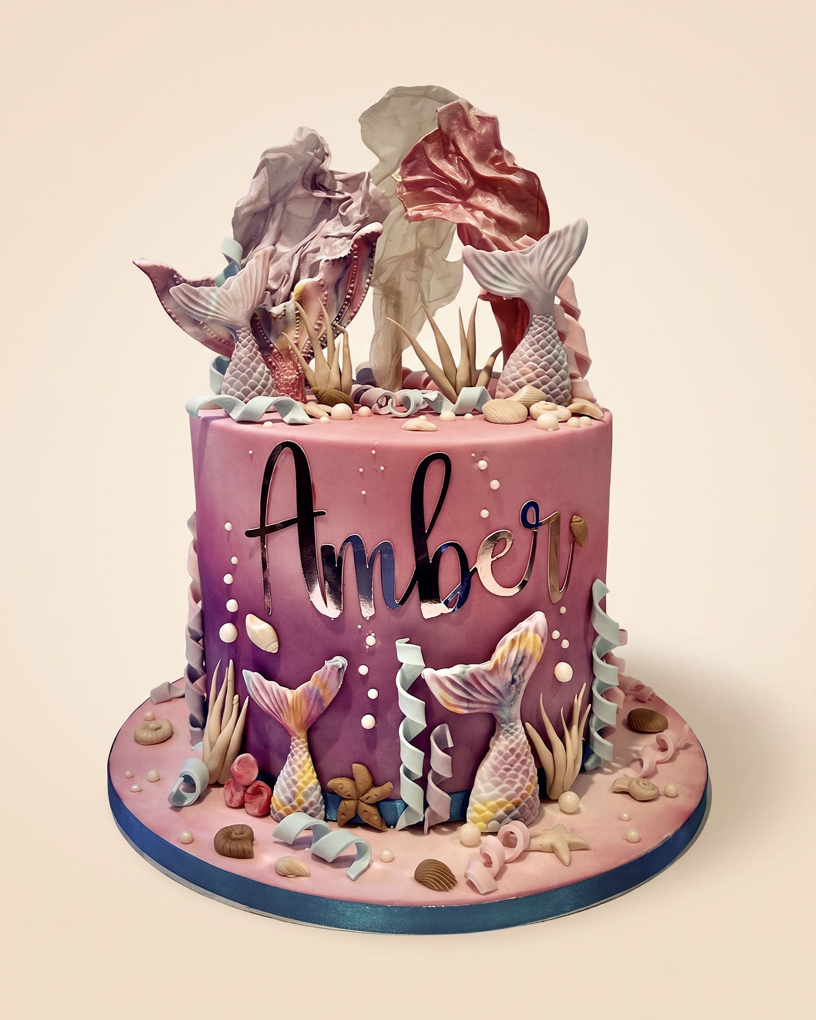 A custom mermaid-themed birthday cake with the name 'Amber' written in black script on the front. The cake features a purple and pink gradient, detailed with fondant mermaid tails, seashells, coral, and sea plants, and is topped with large decorative seashells.