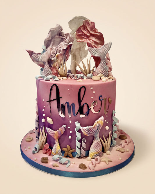 A custom mermaid-themed birthday cake with the name 'Amber' written in black script on the front. The cake features a purple and pink gradient, detailed with fondant mermaid tails, seashells, coral, and sea plants, and is topped with large decorative seashells.
