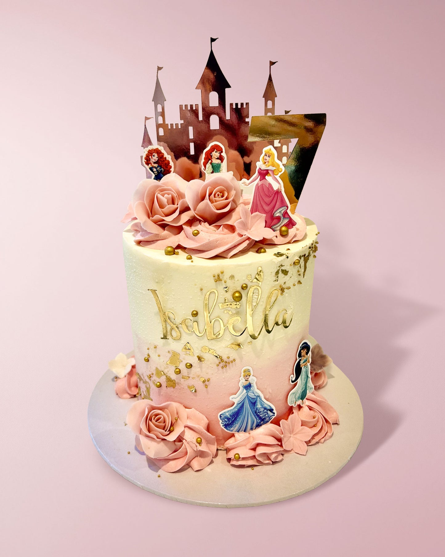 A custom decorated birthday cake with pink frosting roses, gold leaf details, and princess toppers, topped with a castle silhouette on a light pink background.