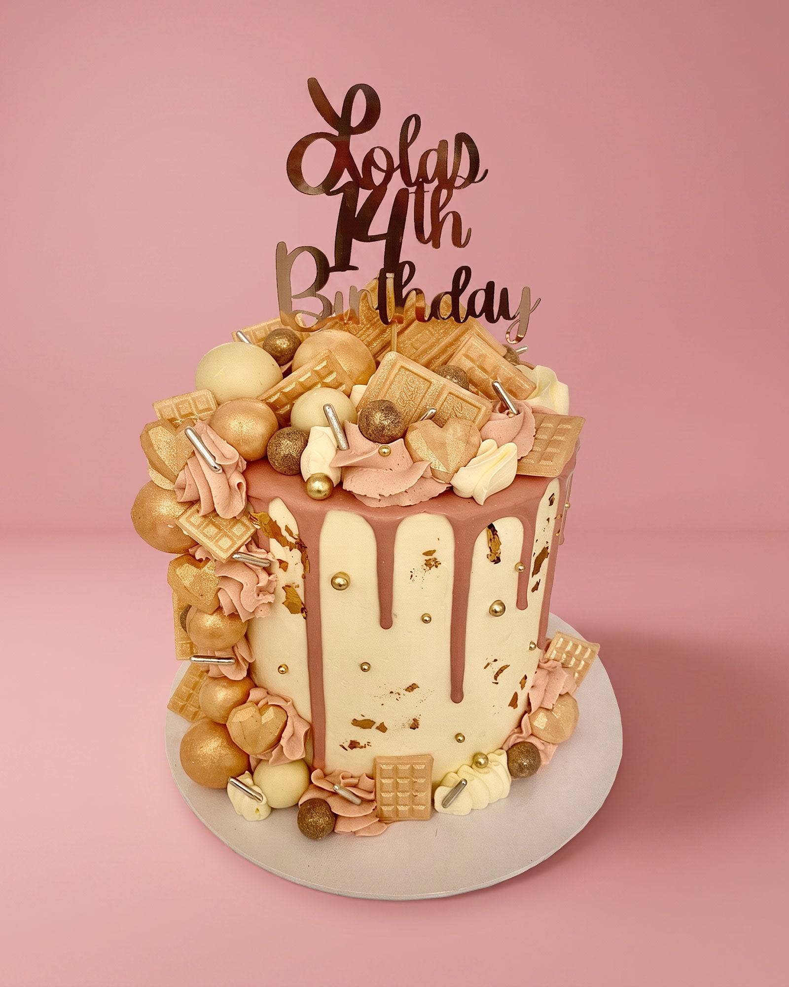 A decorated birthday cake on a white round platter against a solid pink background. The cake has a cream-colored drip frosting base with dusty pink frosting drips, topped with pink and gold chocolate decorations, gummy bears, and a gold 'Lola 14th Birthday' script topper.