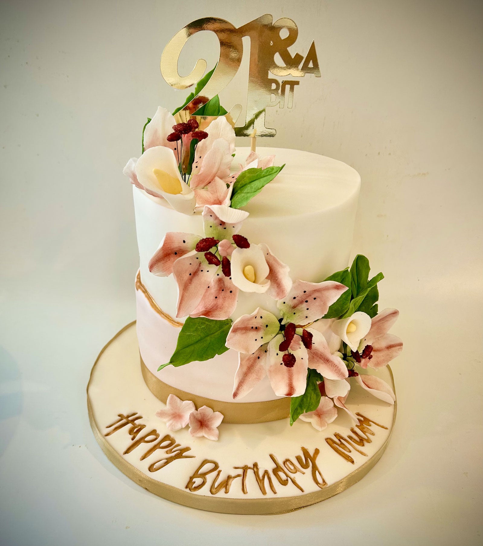 A two-tier white birthday cake decorated with pink lily flowers, green leaves, and a gold 'Mr & Mrs A' topper. The cake sits on a round white board with 'Happy Birthday Mum' written in gold icing, set against a cplain light background.