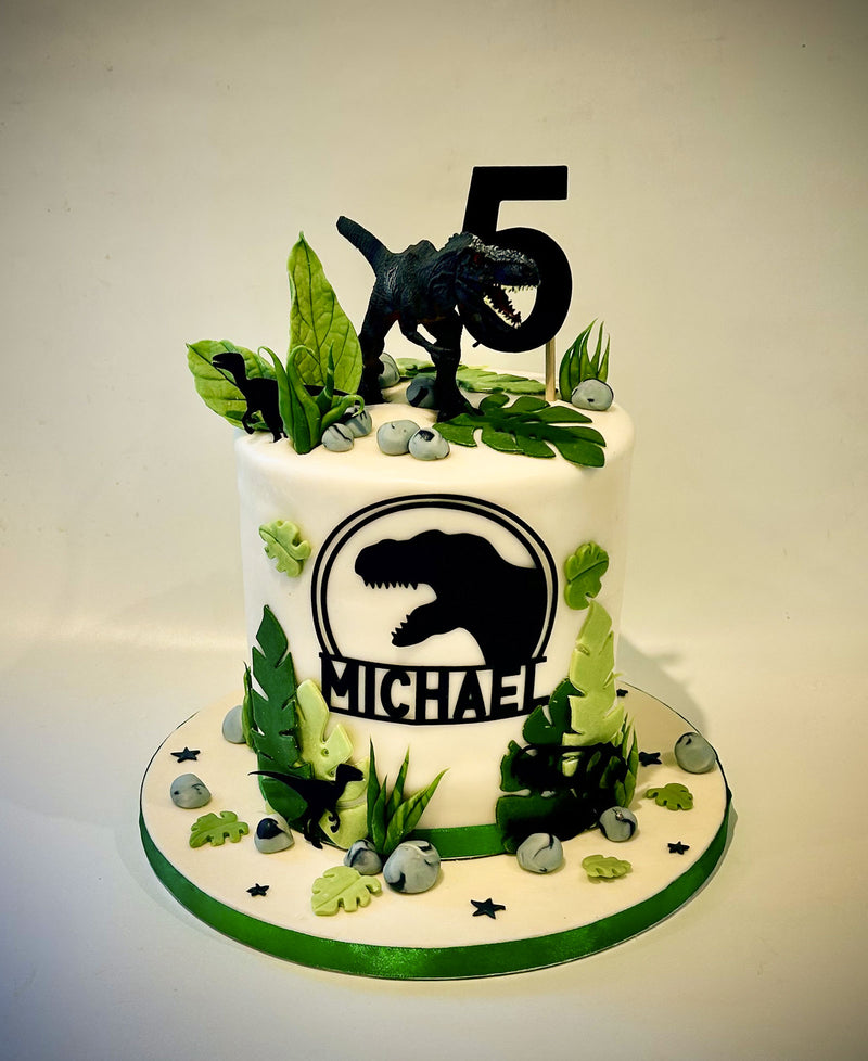 A custom dinosaur-themed birthday cake for a child named Michael, decorated with a black T-Rex silhouette, green leaves, faux rocks, and a black number 5 topper on a white background.