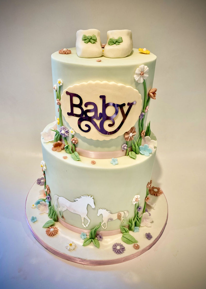 Two-tier baby shower fondant cake with a cream 'Baby' plaque in silver script, white bootee toppers with green bows, floral accents and white horse silhouettes, handmade by Daisy Mud Pie in Brentwood, Essex