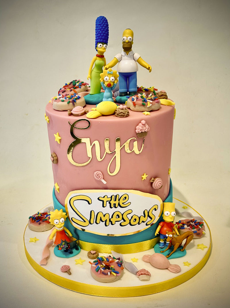 A custom pink and teal tiered cake decorated with The Simpsons character figurines, including Bart, Lisa, and Maggie, along with donut toppers and the name 'Enya' in gold script.