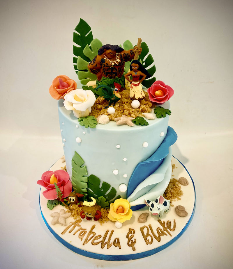 A custom tropical-themed birthday cake for a baby shower, decorated with fondant, coconut shell pieces, green leaves, and two small Polynesian figure toppers. The cake is personalized with the names 'Isabella & Blake' written in gold icing on the base.