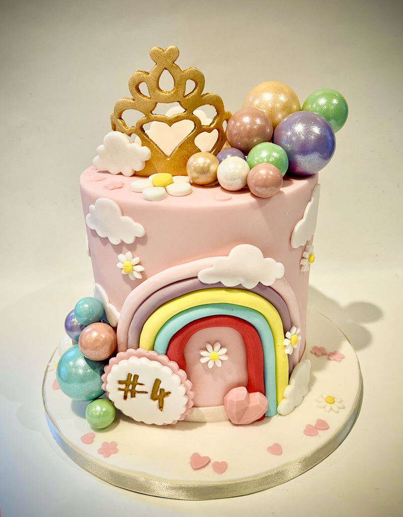 Pastel pink rainbow fairy tale castle fondant cake with gold tiara, colourful fondant clouds, flowers and metallic sphere decorations, handmade by Daisy Mud Pie in Brentwood, Essex