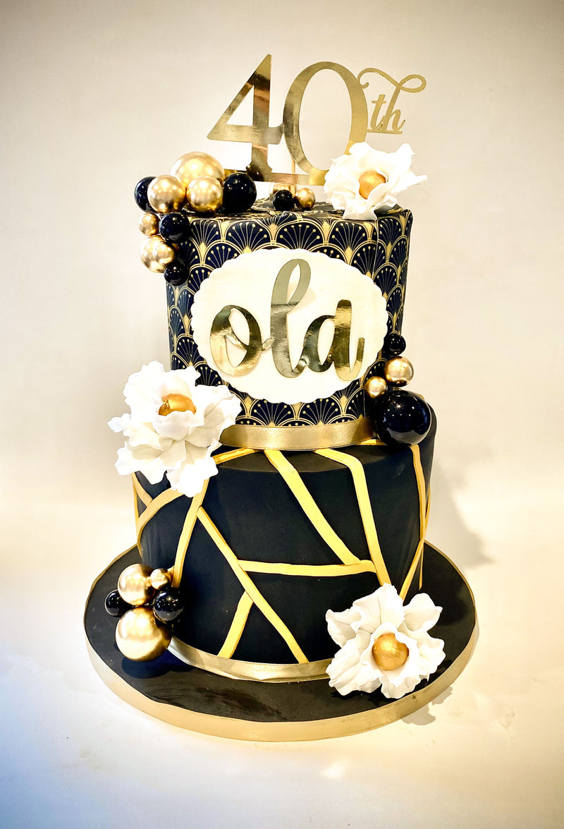 A two-tier black and gold anniversary cake decorated with white flowers, gold and black beads, and a gold '40th' topper, set against a plain light background.