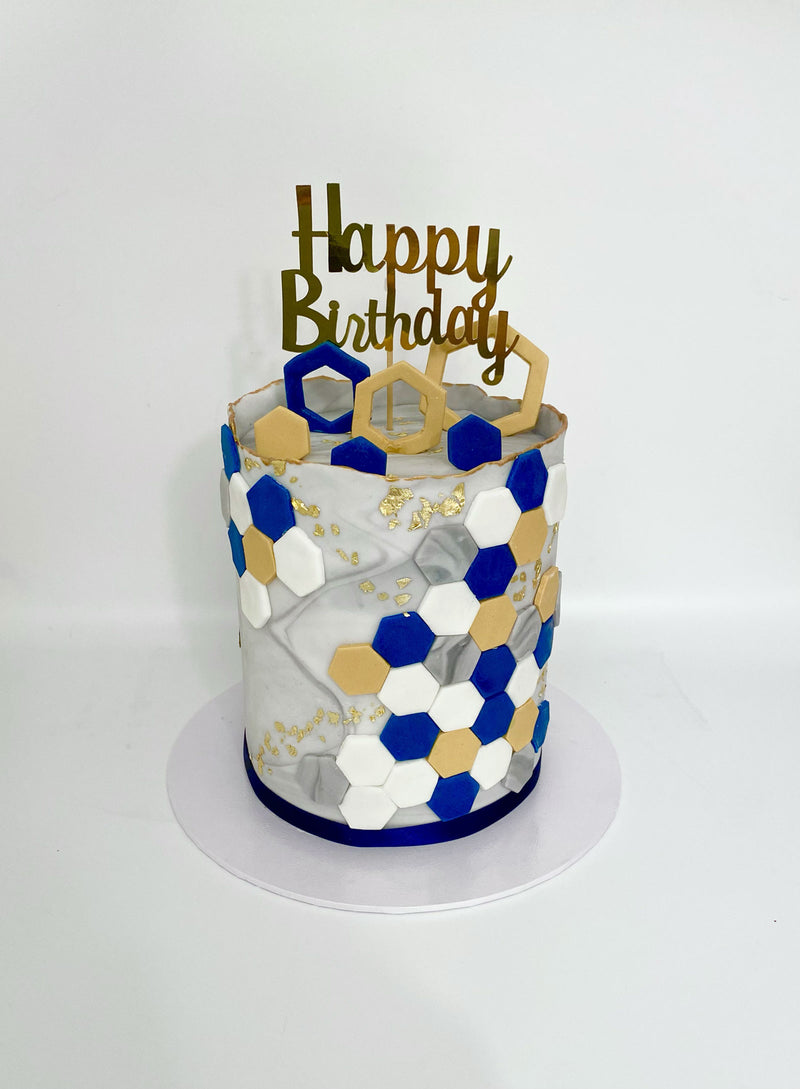 A cylindrical birthday cake decorated with a white and gray marble pattern, blue, white, and gold hexagonal tiles, a gold 'Happy Birthday' topper, and a white round cake board, photographed against a plain white background.