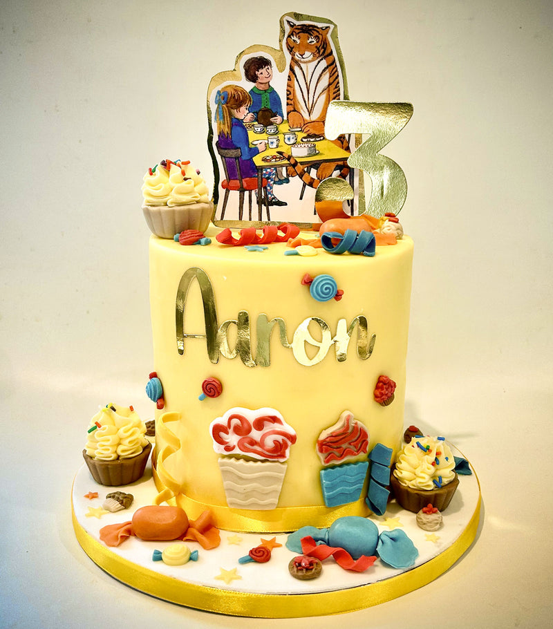 A custom 3-year-old birthday cake for a boy named Aaron, decorated with yellow fondant, gold lettering, a gold number 3 topper featuring a boy and a tiger, and various colorful candy decorations.