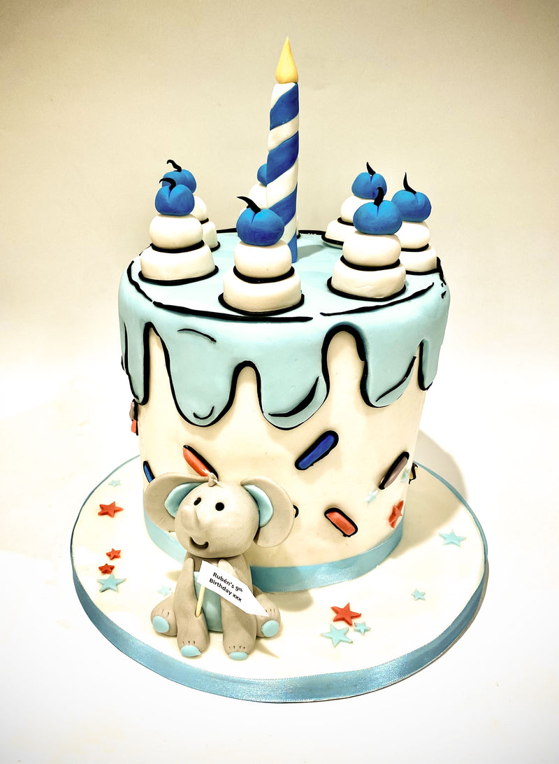 A decorated birthday cake with light blue and white frosting, blue and white cupcake toppers, a tall blue-and-white striped candle, and a small fondant elephant figurine on the front, set on a round matching cake board against a plain light background.