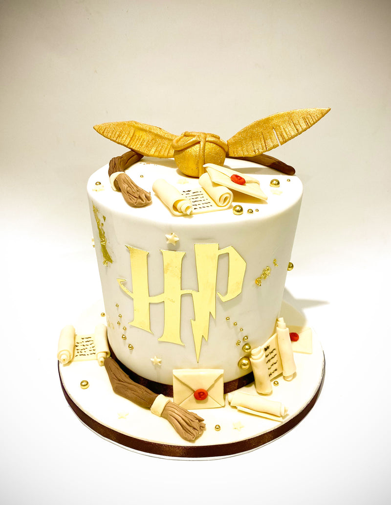 Harry Potter themed fondant cake with gold and beige HP logo, golden snitch, scroll and envelope details, and a broomstick design, handmade by Daisy Mud Pie in Brentwood, Essex