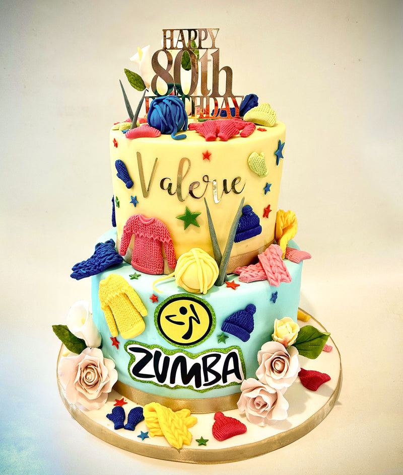 A custom two-tiered Zumba-themed birthday cake for Valerie. The top tier is pale yellow with 'Valerie' written in gold script, decorated with fondant balls, dance shoes, and a 'HAPPY 80th Birthday' topper.