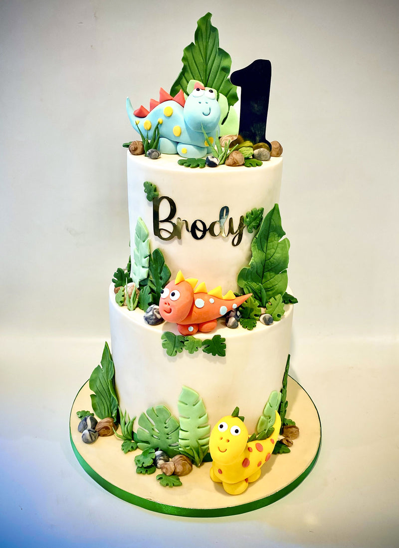 A two-tier white cake decorated with colorful dinosaur figurines, green leaves, faux rocks, and a black number 1 topper. The cake is personalized with the name 'Brody' in black script on the front of the upper tier.