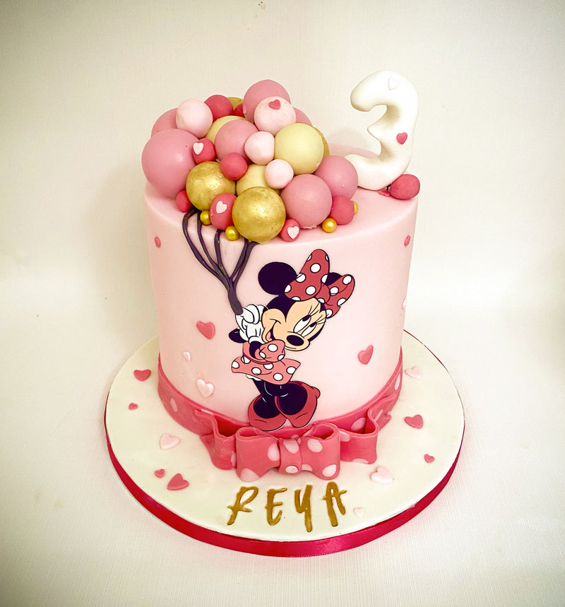 Pink Minnie Mouse fondant birthday cake with Minnie Mouse graphics, pink and gold balloon shapes and a pink fondant bow, handmade by Daisy Mud Pie in Brentwood, Essex