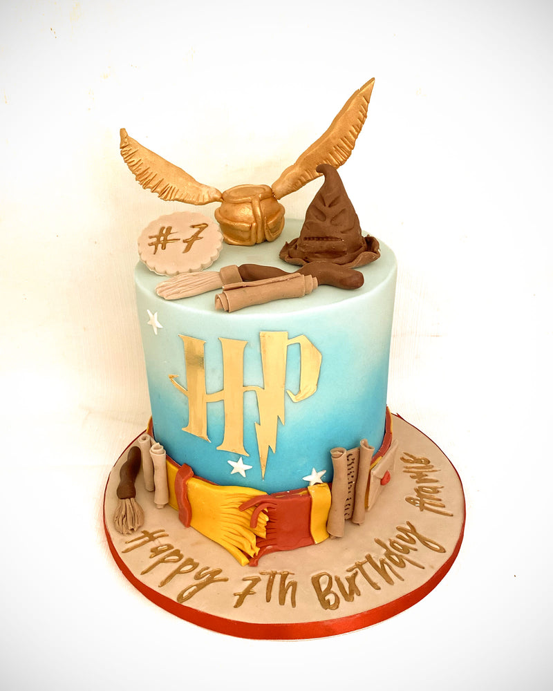 Wizard-themed fondant cake with blue gradient background, gold lettering, golden snitch detailing, wizard hat and wand toppers, handmade by Daisy Mud Pie in Brentwood, Essex