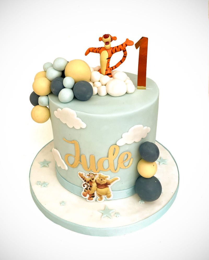 A custom 1st birthday cake decorated with a light blue fondant finish, featuring white clouds, gold script spelling 'Jude', Winnie the Pooh and Tigger figurines, a gold number 1 candle, and decorative pastel balloons on a white cake board.