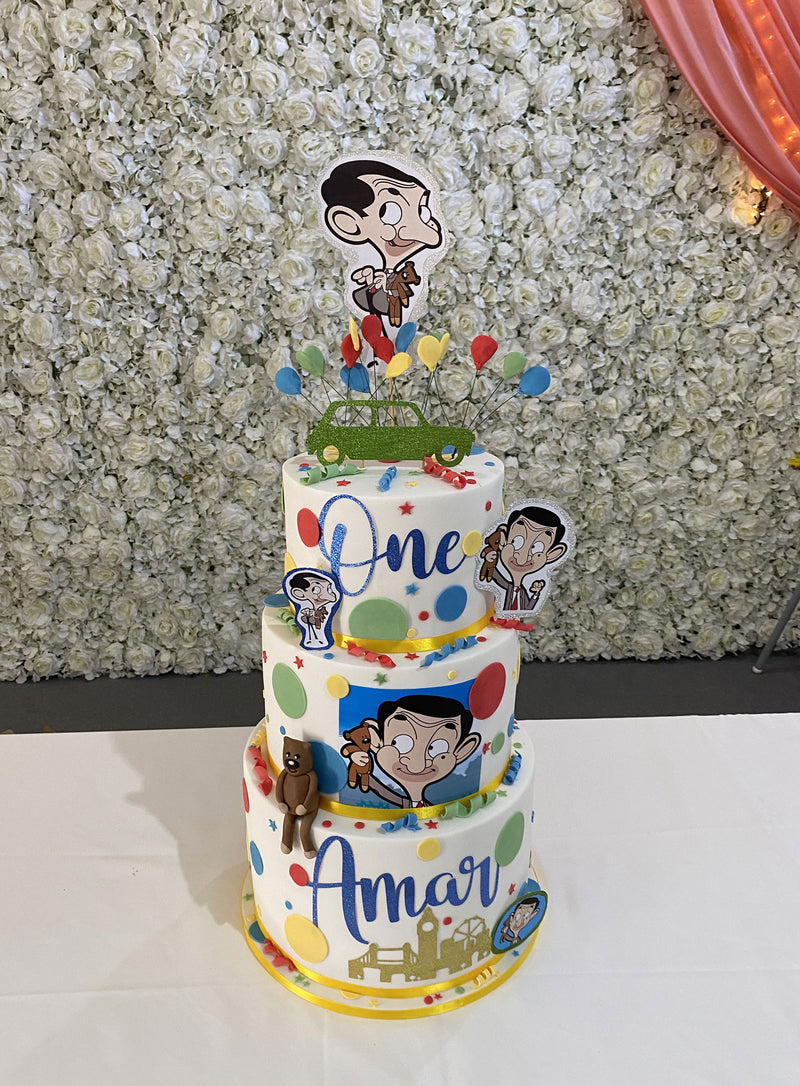 A three-tier custom birthday cake decorated with Mr. Bean cartoon graphics, colorful polka dots, and the names 'One' and 'Amar'.