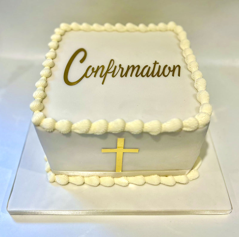 A square white cake decorated with piped cream frosting borders, a gold cross on the front, and the word 'Confirmation' in gold script on txop, displayed on a clear acrylic stand.