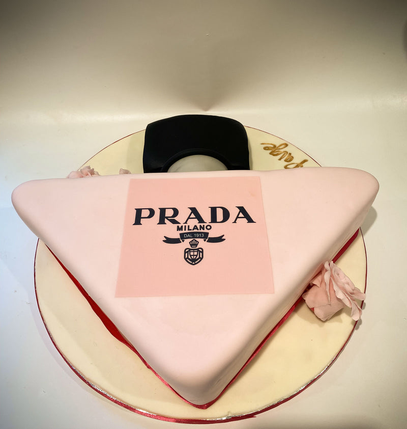 A triangular Prada Milano cake with pale pink fondant, featuring a central pink square with the black Prada logo and a black-and-white circular topper behind it, displayed on a cream-colored round cake board.