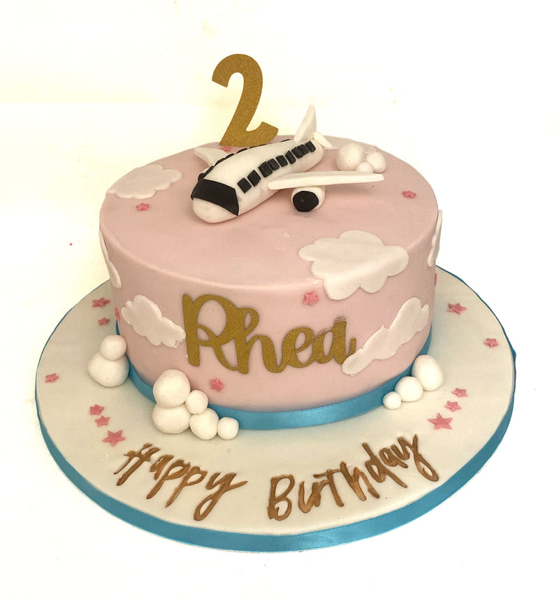 Pink fondant birthday cake with white cloud detailing, airplane topper, gold number candle and gold script cake board, handmade by Daisy Mud Pie in Brentwood, Essex